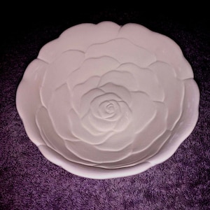 May include: A white, ceramic bowl with a rose design. The bowl is round with a scalloped edge and a raised floral pattern. The bowl is sitting on a purple surface.