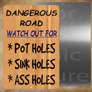 May include: A rectangular metal sign with a wood grain background. The sign reads "DANGEROUS ROAD WATCH OUT FOR * POT HOLES * SINK HOLES * ASS HOLES" in black and blue text. The sign has two holes at the top for mounting.
