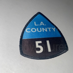 May include: A triangular patch with a blue top section and a black bottom section. The text "L.A. COUNTY" is in white on the blue section, and the number "51" is in white on the black section. The patch has a black border.