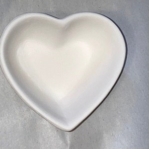May include: A heart-shaped, off-white ceramic dish. The dish has a smooth, glossy finish and a shallow interior. The heart shape is clearly defined, with rounded edges and a symmetrical design. This decorative dish is suitable for small items.