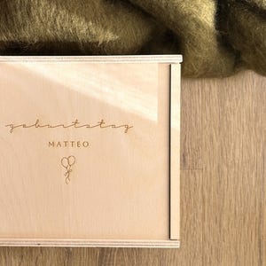 Könnte beinhalten: Eine hellfarbene Holzkiste mit dem Wort "geburtstag" und dem Namen "MATTEO" auf der Vorderseite eingraviert. Eine kleine Illustration von Luftballons ist ebenfalls vorhanden. Die Kiste steht vor einem Hintergrund aus olivgrünem Stoff und einer Holzoberfläche.