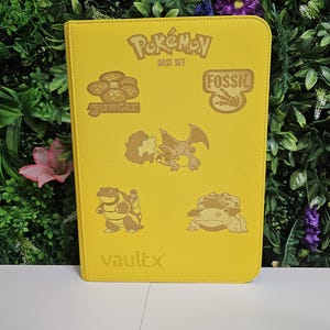 May include: Yellow Vault X Pokémon card binder with embossed Pokémon characters and logos. Features the text "Pokémon Base Set", "Jungle", and "Fossil". The binder is on a white surface with a green and purple floral background.