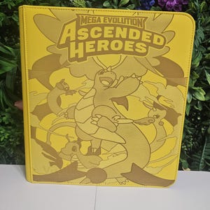 May include: Yellow card binder with the text "MEGA EVOLUTION ASCENDED HEROES" and cartoon illustrations of creatures. The binder is a solid yellow colour with a stitched border.