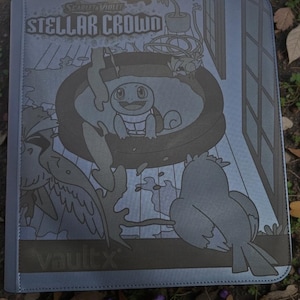 Può includere: Un raccoglitore di carte blu con la scritta "STELLAR CROWD" e "Vault X". Il raccoglitore presenta un'illustrazione a cartoni animati di una tartaruga e altri personaggi. Il raccoglitore è appoggiato su un letto di foglie e piccoli fiori viola.