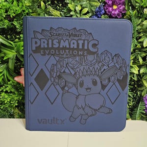May include: A navy blue Vault X binder featuring the text "SCARLET & VIOLET PRISMATIC EVOLUTIONS" and an illustration of Eevee. The binder has a diamond pattern and is surrounded by greenery.