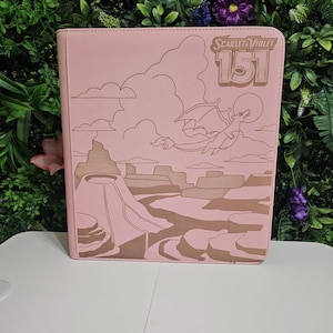 May include: A pink binder with a brown illustration of a dragon, clouds, and a canyon landscape. The text "Scarlet & Violet 151" is printed on the upper right corner. The binder is on a white surface with a green and purple floral background.