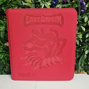 Puede incluir: Un archivador rojo Vault X con el diseño "Lost Origin" de la serie "Sword & Shield". La portada muestra una ilustración detallada de una criatura con apéndices puntiagudos. El archivador está diseñado para guardar cartas coleccionables.