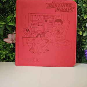 May include: A red Vault X binder featuring a detailed illustration of a person in a chair with a pet, a desk, and a bookshelf. The top right corner displays the text "Scarlet & Violet DESTINED RIVALS". The Vault X logo is at the bottom.