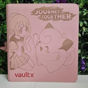 Puede incluir: Carpeta rosa Vault X con una ilustración de dibujos animados de una niña y un Clefairy. La carpeta tiene el texto "Scarlet & Violet Journey Together" y el logotipo de Vault X. Diseñada para cartas coleccionables.