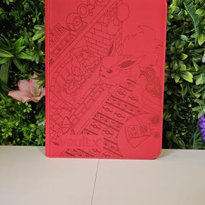 Puede incluir: Una funda roja para tableta con un diseño grabado detallado que presenta un personaje de dibujos animados, una chimenea y un libro. La palabra "vaultx" está impresa en la esquina inferior izquierda.