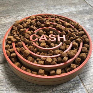 May include: A circular slow feeder dog bowl with a salmon-colored rim and interior design. The bowl is filled with brown kibble and features the word "CASH" in the center. The design is intended to slow down eating.