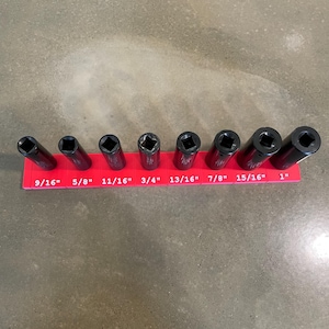 May include: A set of black socket wrenches of varying sizes, arranged on a red holder. The sizes are labeled in inches, ranging from 9/16" to 1". The tools are designed for mechanical work and are set against a neutral background.
