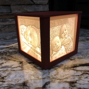 May include: A lighted, cube-shaped lamp with a dark brown frame. The lamp's sides display etched images of children. The lamp is plugged into an electrical outlet. The lamp is sitting on a marble surface.