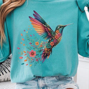 May include: A seafoam green long-sleeve sweatshirt featuring a vibrant, multi-colored hummingbird graphic. The bird is depicted with intricate floral patterns on its wings and body, with colorful leaves and flowers trailing behind it in a dynamic motion.