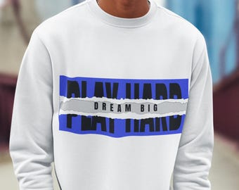 Könnte beinhalten: Weißes Sweatshirt mit dem Text "PLAY HARD DREAM BIG" in einem blau-grauen Design auf der Brust. Das Sweatshirt hat lange Ärmel und wird mit einer marineblauen Hose kombiniert. Der Hintergrund zeigt eine Stadtlandschaft und eine rote Brücke.