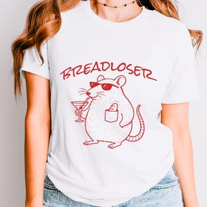 May include: White t-shirt with a red graphic of a rat wearing sunglasses and holding a cocktail. The text "BREADLOSER" is above the rat. The rat has a pocket with a small object inside.