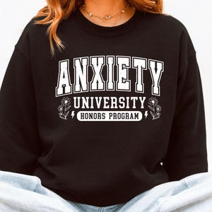 Op de afbeelding: Zwart sweatshirt met de witte tekst "ANXIETY UNIVERSITY HONORS PROGRAM". De sweatshirt heeft een ronde hals en lange mouwen. De tekst staat in het midden op de voorkant.