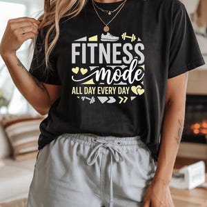 May include: Black t-shirt with the text "FITNESS mode ALL DAY EVERY DAY" in white and yellow. The design includes a shoe, dumbbell, and heart icons. The person is wearing gray shorts.