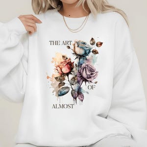 May include: White sweatshirt with a watercolour floral design. The design features roses in red, blue, and purple. The text "THE ART OF ALMOST" is printed above and below the floral arrangement.