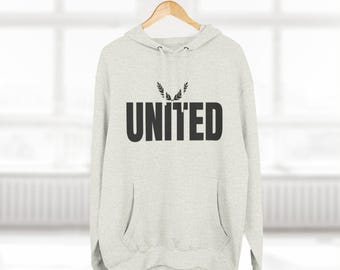 May include: A light grey hoodie featuring the word "UNITED" in bold black lettering. Above the text is a small black sprig design. The hoodie has a front pocket and a hood, and hangs from a clothes rail.