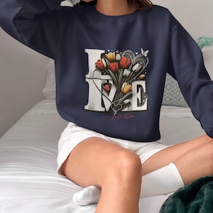 May include: Navy blue sweatshirt featuring the word "LOVE" in white block letters. A bouquet of red, yellow, and white tulips is in the centre. The words "in full bloom" are written in cursive below the "E".