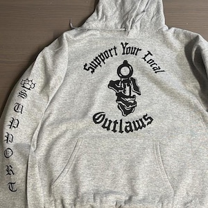 May include: A gray hoodie with a front pocket and a hood. The back of the hoodie features the text "Support Your Local Outlaws" above a graphic. The sleeves have the word "Outlaws" and a brass knuckle graphic.