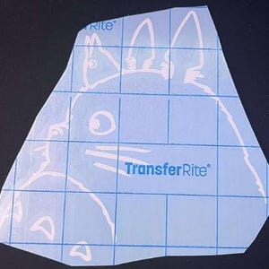 Puede incluir: Calcomanía de vinilo blanco de un personaje de dibujos animados, con un cuerpo grande, ojos grandes y orejas puntiagudas. La calcomanía está sobre una hoja de cuadrícula azul con el texto "TransferRite".