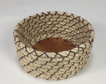 May include: A small, round woven basket with a brown leather base. The basket is made of natural fiber rope and has a decorative brown stitching detail.