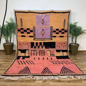 May include: A rectangular area rug featuring a color-blocked design in shades of orange, pink, and purple. The rug displays black geometric patterns, including diamonds, checkered stripes, and triangles, with a fringed edge along the bottom.