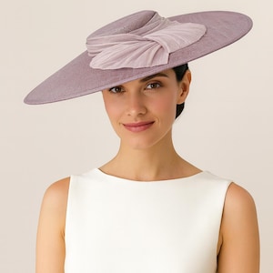 Puede incluir: Una mujer lleva un sombrero lavanda con ala ancha y un lazo decorativo. El sombrero está hecho de una tela texturizada y tiene un aspecto suave y fluido. La mujer lleva una camiseta sin mangas blanca.