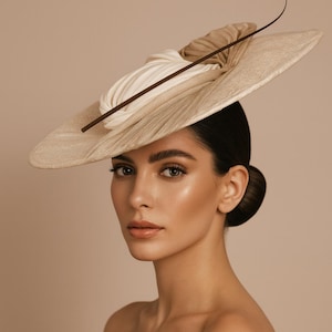 May include: A beige wide-brimmed hat with a circular design, adorned with cream and tan fabric detailing. A dark brown accent piece crosses the hat, with a thin, curved element extending upwards. This elegant hat is a fashion accessory, suitable for special occasions.
