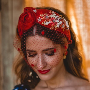 Puede incluir: Una diadema roja con una gran rosa de tela, intrincados abalorios florales y un velo negro tipo jaula con pequeños lunares rojos. La diadema se combina con un top azul marino de un solo hombro con un estampado de lunares blancos.