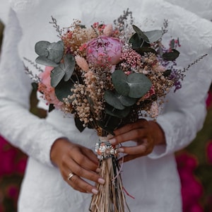 May include: A bride holds a bouquet of dried flowers, including pink peonies, eucalyptus, and dried grasses. The bouquet is tied with a brown ribbon and has a small, decorative charm.