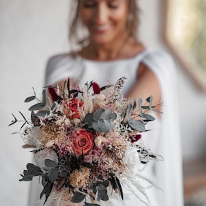 May include: A bridal bouquet with dried flowers in shades of pink, white, and beige. The bouquet features eucalyptus leaves, dried grasses, and small pink roses.