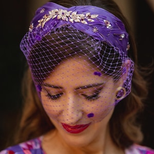 May include: A wide purple headband featuring a textured feather-like surface adorned with gold-toned metal floral embellishments and small purple crystals, paired with a matching purple birdcage veil with scattered purple velvet dots.