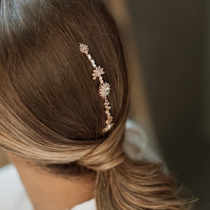 Pode incluir: Um pente de cabelo dourado com pequenos detalhes de pérolas e desenhos florais. O pente é usado no cabelo de uma mulher, que está preso em um rabo de cavalo baixo.
