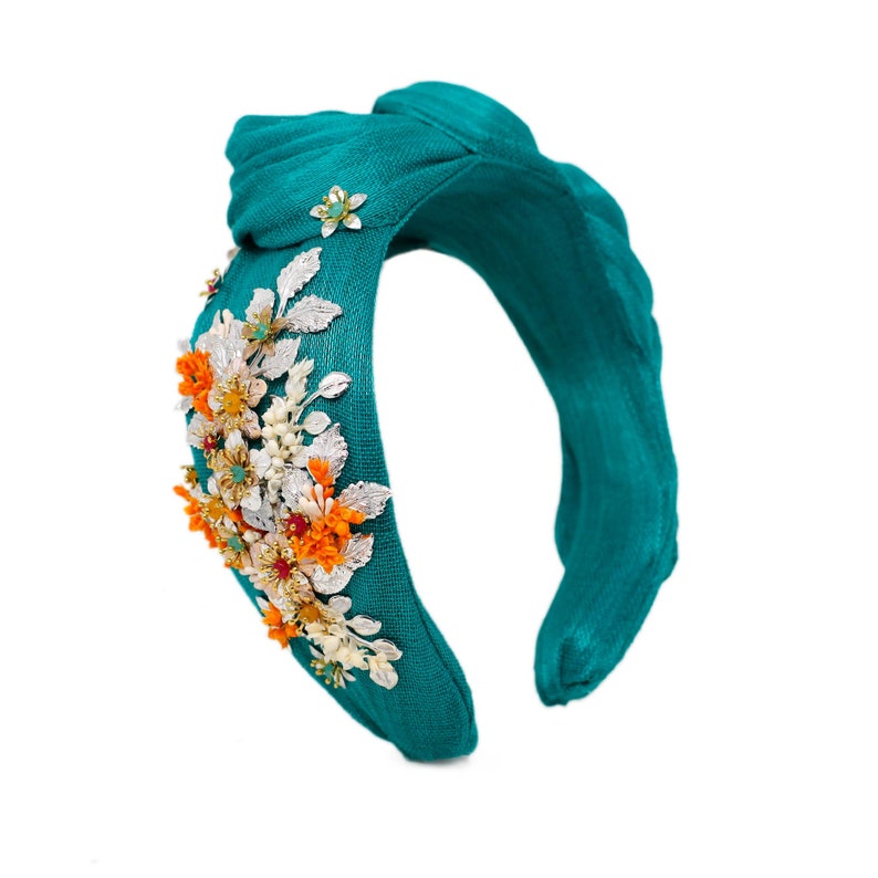 May include: A teal knotted headband featuring a textured fabric surface adorned with a cluster of metallic silver leaves, bright orange floral accents, and small embedded green crystals arranged along the side of the band.