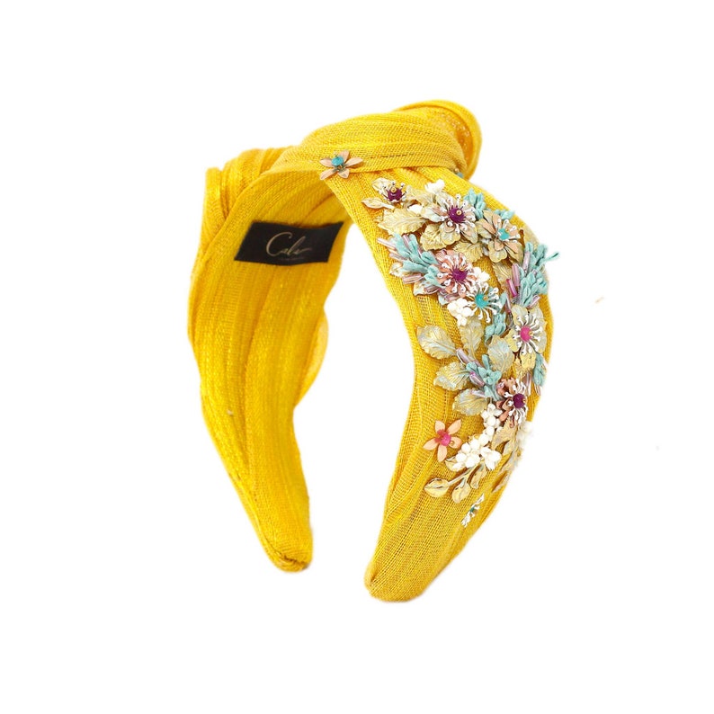 May include: A bright yellow knotted headband featuring a textured fabric finish and a black label with the text Cala. The side is adorned with a cluster of metallic floral embellishments in shades of gold, light blue, white, and pink.
