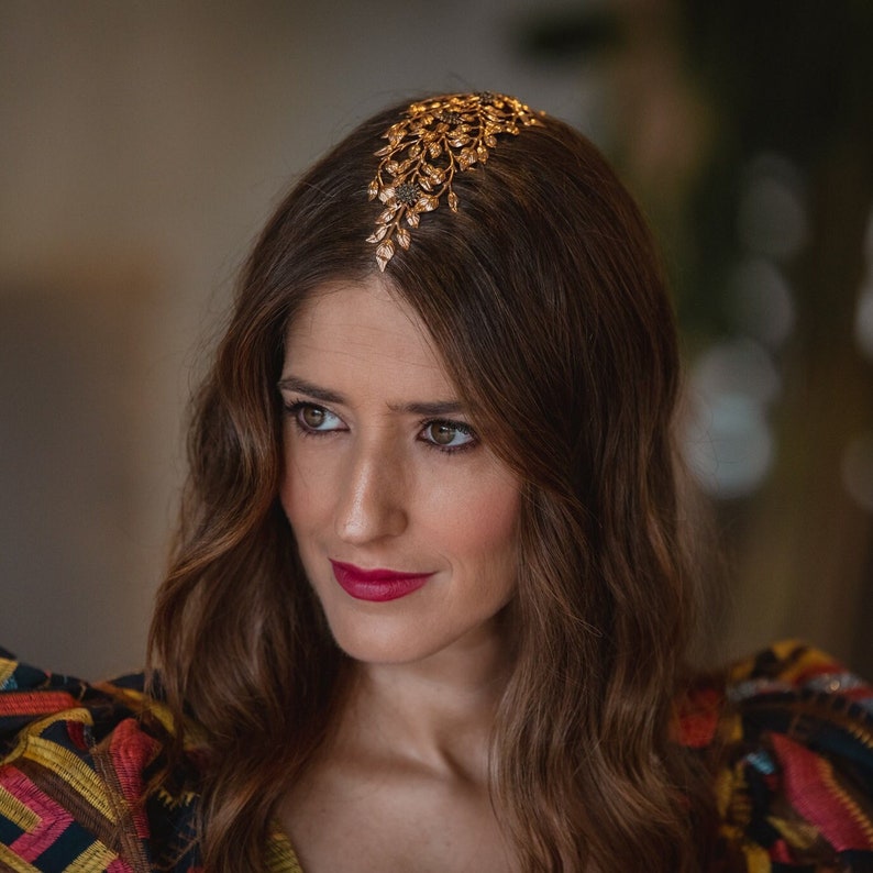 May include: A woman with long brown hair wearing a gold hairpiece with a leaf design. She is wearing a colorful patterned shirt.