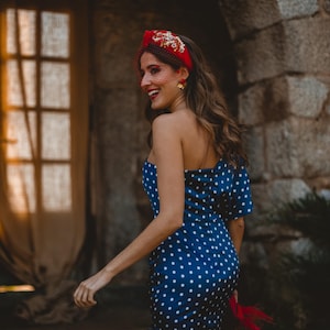 May include: A navy blue one-shoulder dress featuring a white polka dot pattern and a side slit. The ensemble includes a red headband adorned with gold floral embellishments and a fine red netting veil, paired with gold flower-shaped earrings.