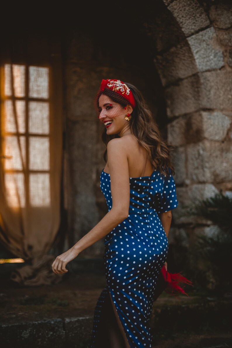 May include: A navy blue one-shoulder dress featuring a white polka dot pattern and a side slit. The ensemble includes a red headband adorned with gold floral embellishments and a fine red netting veil, paired with gold flower-shaped earrings.