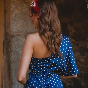 May include: A navy blue one-shoulder dress featuring a white polka dot pattern, a fitted bodice with a central back zipper, and a single flared sleeve. The ensemble is accessorized with a red headband adorned with floral details and a mesh veil.