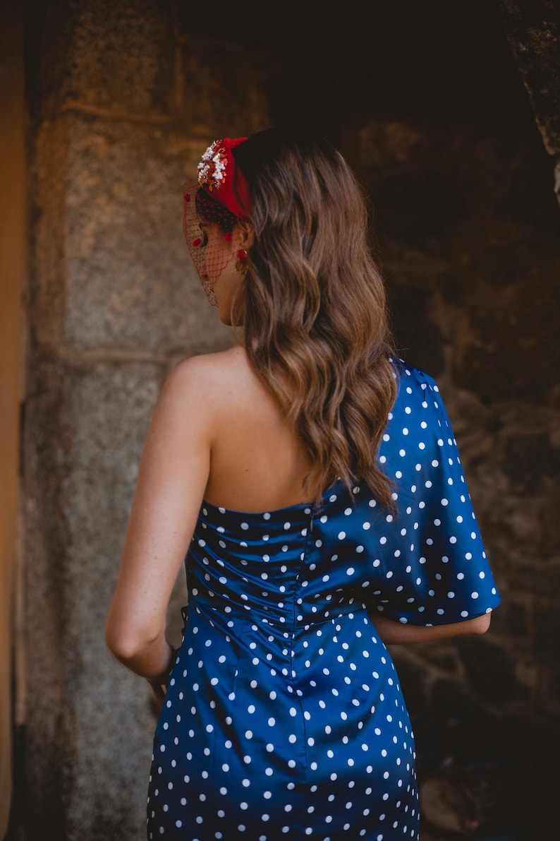 May include: A navy blue one-shoulder dress featuring a white polka dot pattern, a fitted bodice with a central back zipper, and a single flared sleeve. The ensemble is accessorized with a red headband adorned with floral details and a mesh veil.