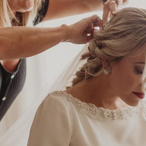 Könnte beinhalten: Eine Braut wird mit einem zarten Haarschmuck frisiert. Sie trägt ein weißes Kleid mit einem perlenbesetzten Ausschnitt und einem Schleier. Das Bild zeigt die Vorbereitung auf eine Hochzeit, wobei die Details der Braut im Mittelpunkt stehen.