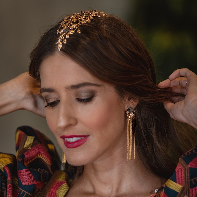 May include: A woman wearing a gold headband with a floral design and matching gold earrings with a dangling chain. The earrings have a small, dark gray flower-shaped charm.