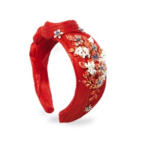 May include: A bright red fabric headband featuring a braided top knot design. The headband is adorned with a cluster of metallic gold floral embellishments, white beaded accents, and small blue stones arranged in a decorative pattern on one side.
