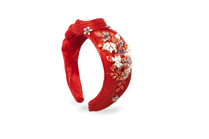 May include: A bright red fabric headband featuring a braided top knot design. The headband is adorned with a cluster of metallic gold floral embellishments, white beaded accents, and small blue stones arranged in a decorative pattern on one side.