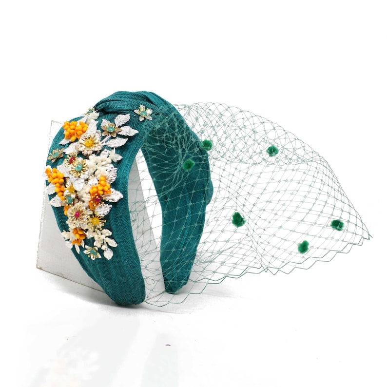 May include: A teal knotted headband featuring a textured fabric surface adorned with a cluster of metallic silver leaves, white flowers, and small yellow berries. A matching teal netting veil with small green pom-poms extends from the side.