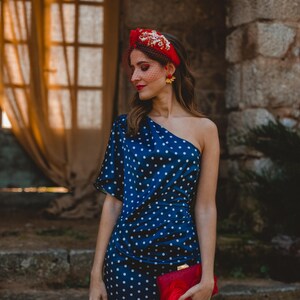 May include: A navy blue one-shoulder dress with white polka dots, paired with a red headband featuring gold floral embellishments and a red veil. A red clutch bag with red feather accents is held in the hand, completing the ensemble.