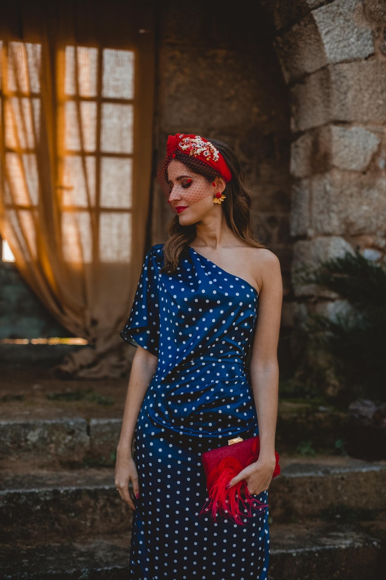 May include: A navy blue one-shoulder dress with white polka dots, paired with a red headband featuring gold floral embellishments and a red veil. A red clutch bag with red feather accents is held in the hand, completing the ensemble.